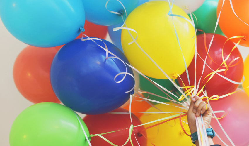 A hand with a blue wristwatch holds a cluster of blue, yellow, green, red, and orange Welcome balloons