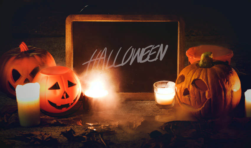 Black chalkboard sign with Halloween written on it surrounded by jack-o-lanterns, candles, and dead leaves