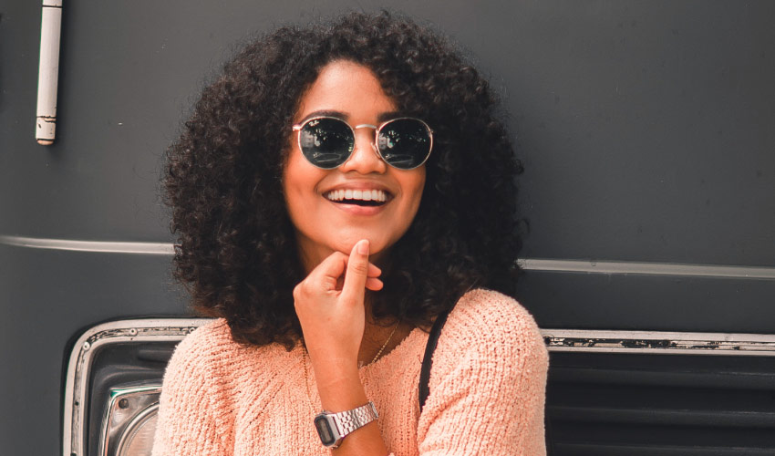 Curly-haired woman wearing sunglasses, peach sweater, and a silver watch smiles with veneers