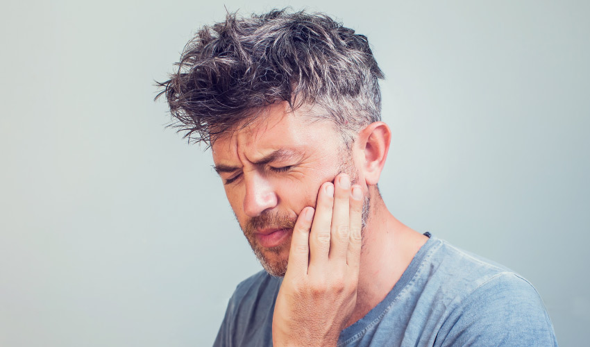 Middle-aged man in a gray shirt winces and touches his cheek due to an emergency toothache