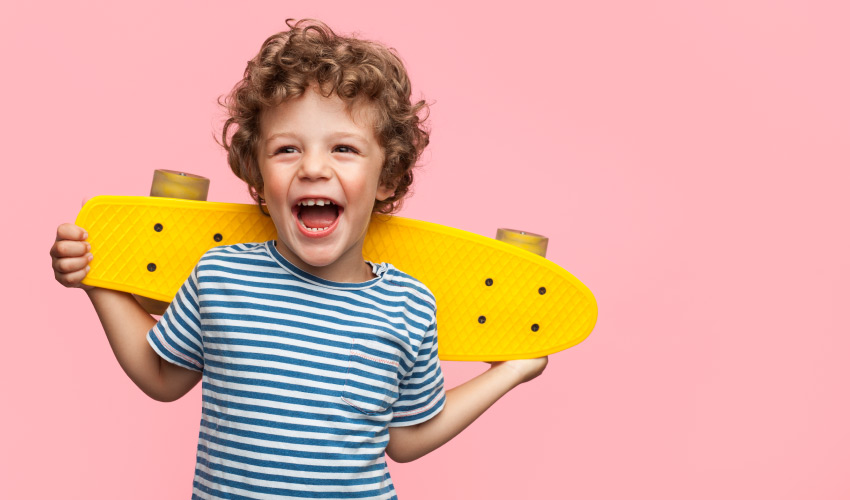 Brunette boy holding a yellow skateboard against a pink wall smiles while laughing