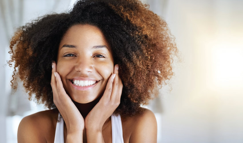 Curly-haired woman with porcelain veneers smiles brightly as she touches the sides of her face