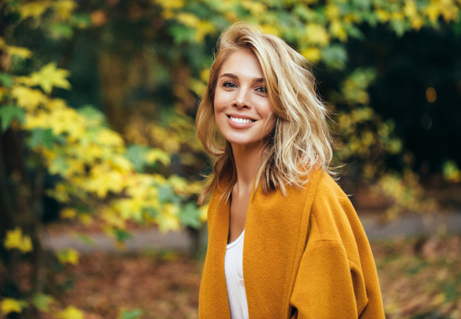 Blonde woman wearing a mustard-colored jacket in the fall smiles with her dental implants