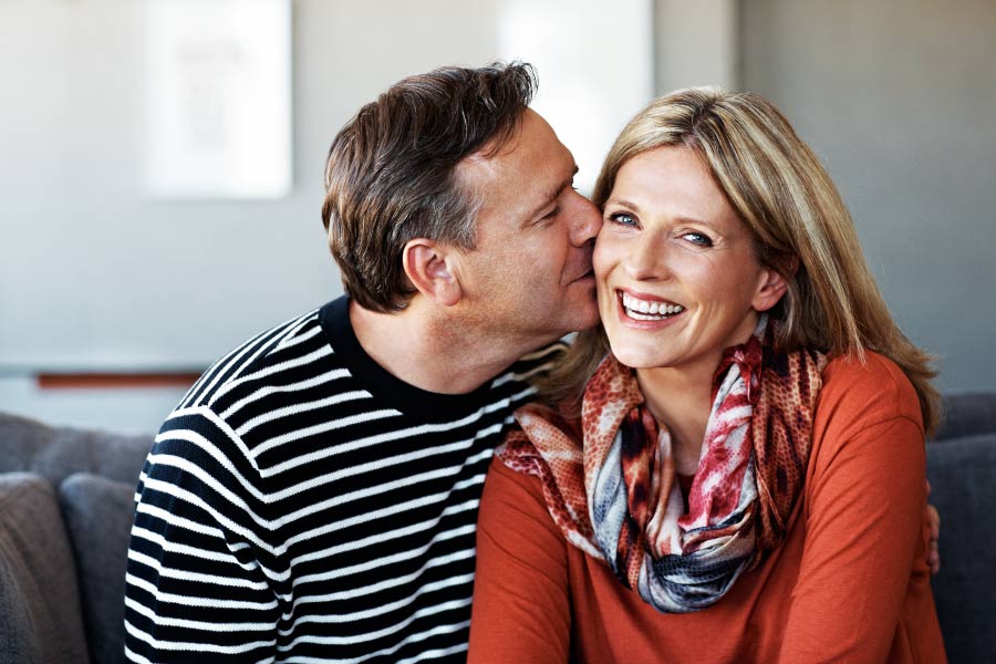 Husband in a striped shirt kisses his wife in a red shirt and scarf on the cheek before receiving sedation dentistry