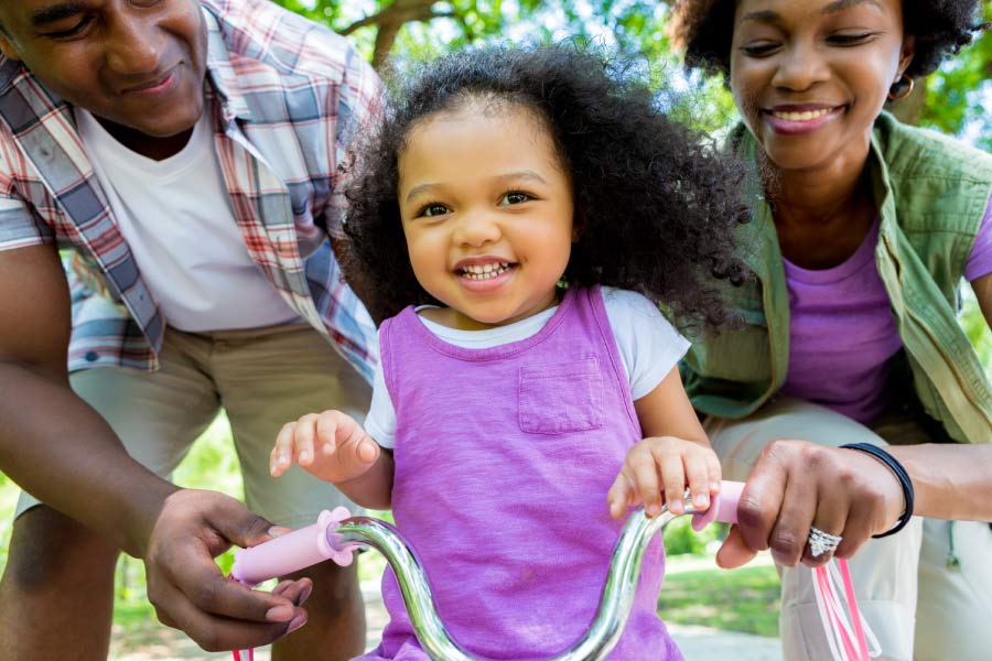 Dad and Mom helping their cute little daughter ride her tricycle.